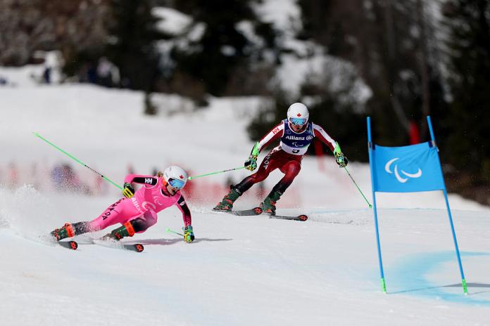 A male Para alpine skier in competition, he is guided by a female skier wearing a pink uniform