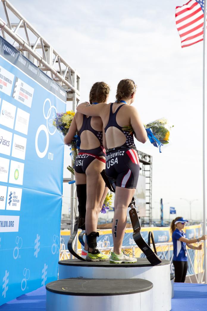 Three Para triathlete women on the podium while the US flag is being raised