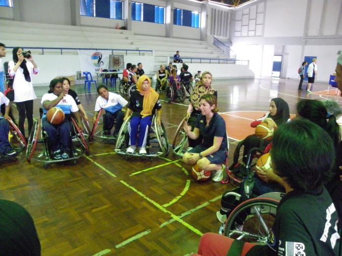 Un entrenador se arrodilla mientras las mujeres en silla de ruedas se reúnen a su alrededor. 