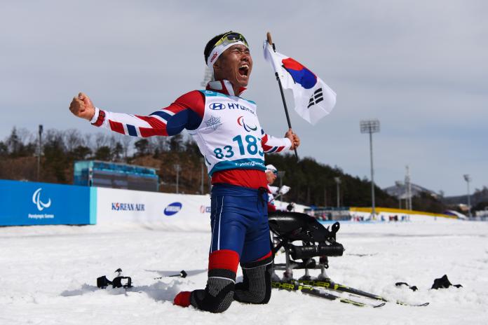 a sit skier celebrates on his knees holding the South Korean flag