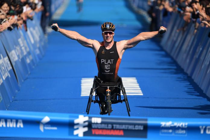male Para triathlete raises his arms in celebration as he breaks the finish line tape