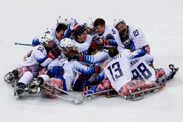 American male ice hockey players celebrate hugigng in a large group on the ice