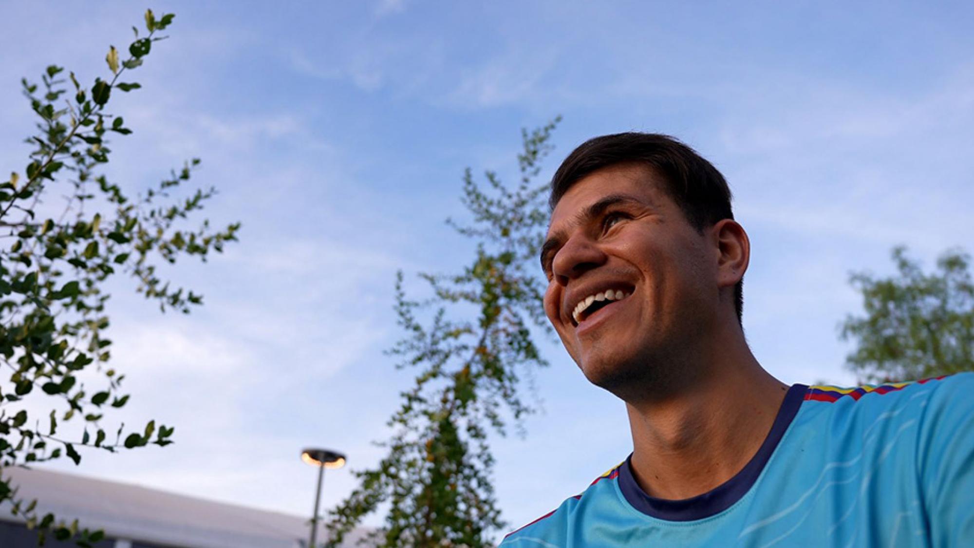 Jhon Gonzalez, a blind football player from Colombia, is standing outdoors wearing a blue uniform, crossing his arms