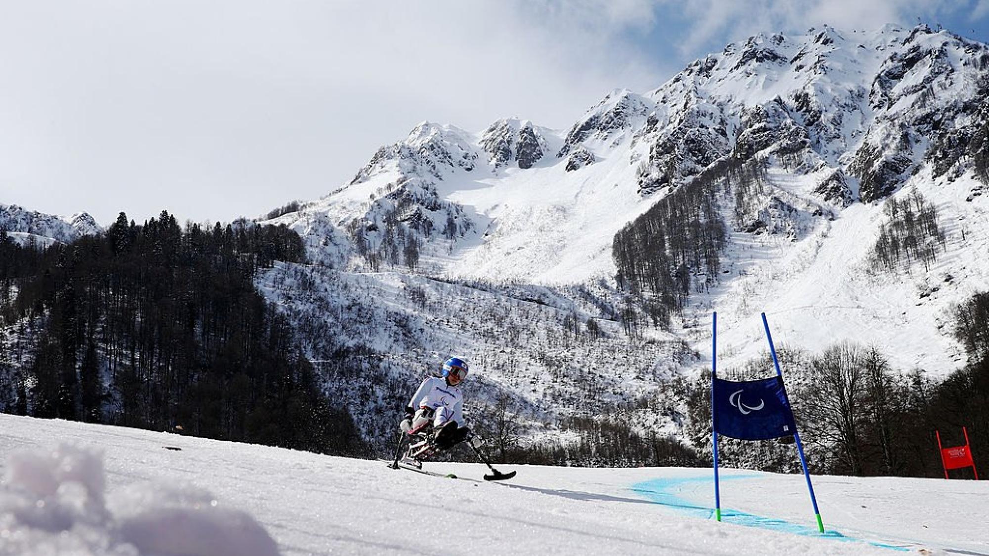 A female sit-skiing athlete is skiing down a slope, with a snow-capped mountain in the background
