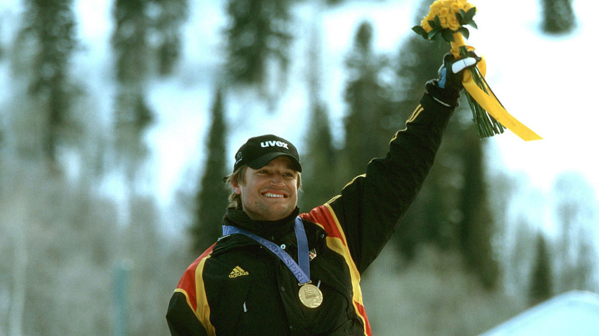 A male athlete wearing a black uniform and a gold medal is celebrating, holding a flower bouquet with his left hand