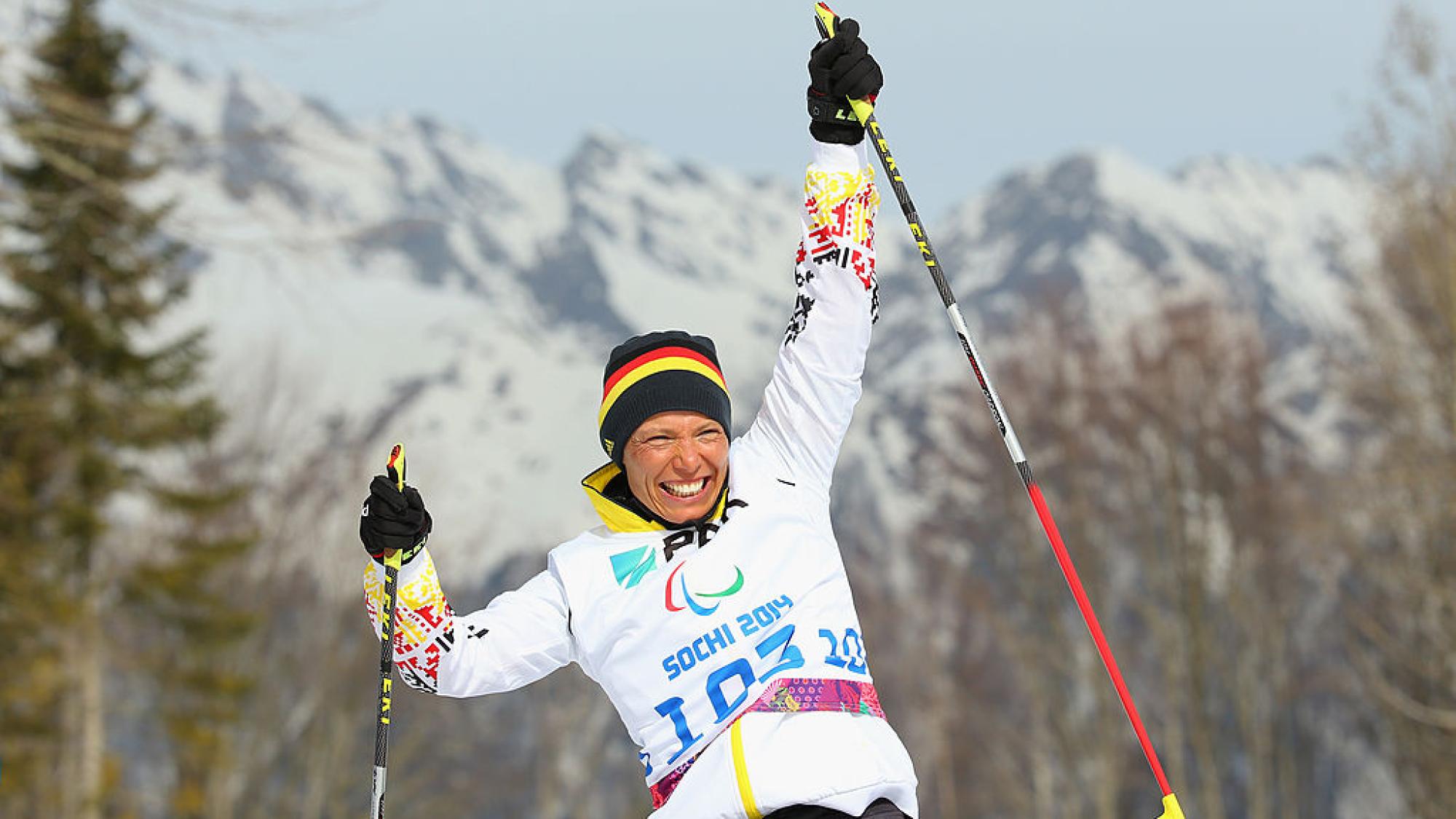 A female athlete holding  two ski poles smiles and raises her arms 