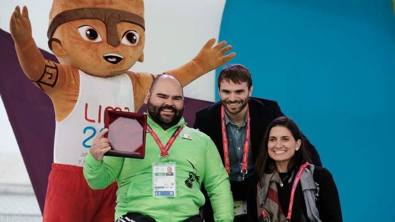 A male powerlifter holds up an award smiling with two other people and a furry mascot standing behind