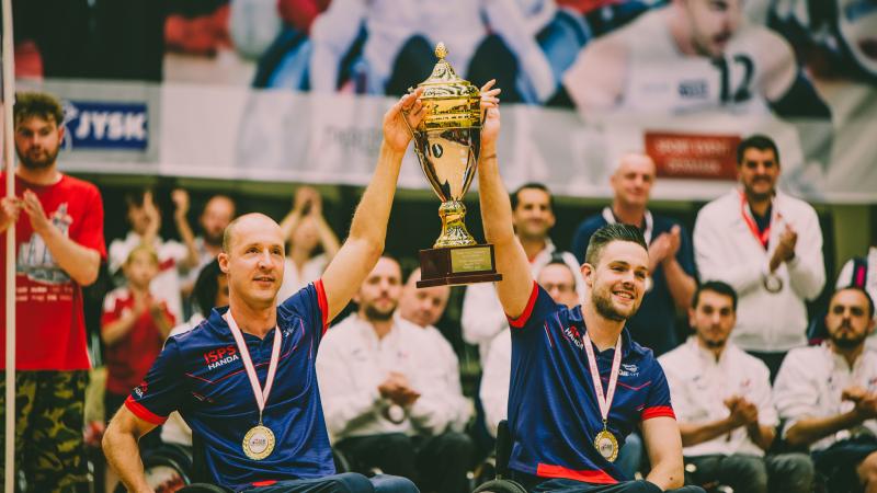 two male wheelchair rugby players with medals around their neck lift a gold cup into the air