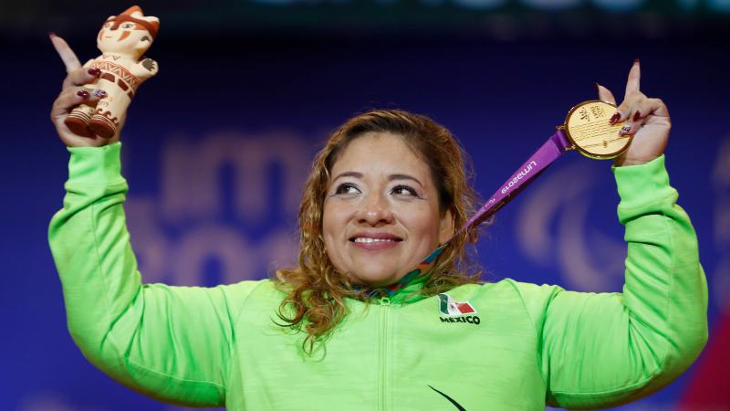 a female powerlifter holds up her gold medal and points her fingers to the sky