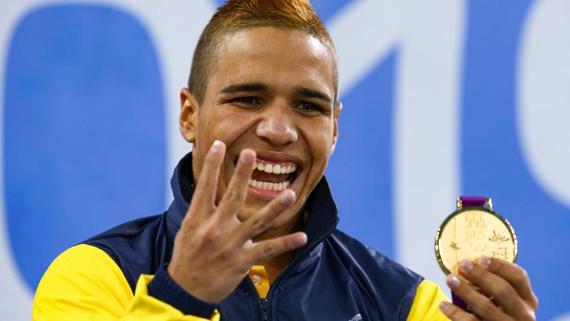 a male Para swimmer holds up four fingers and his gold medal