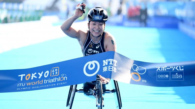 a female Para triathlete in a wheelchair raises her arm as she breaks the finish line tape