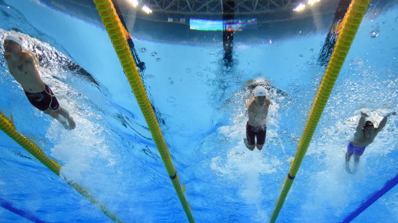 three male Para swimmers with no arms swimming breaststroke