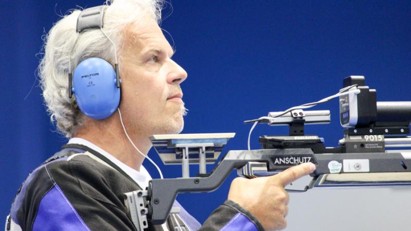 Male vision impaired rifle shooting athlete looks on with noise canceling ear protectors on