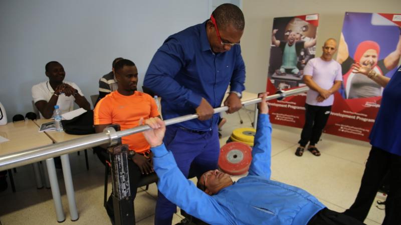 A man on a bench lifting a bar with the support of another man observed by five people