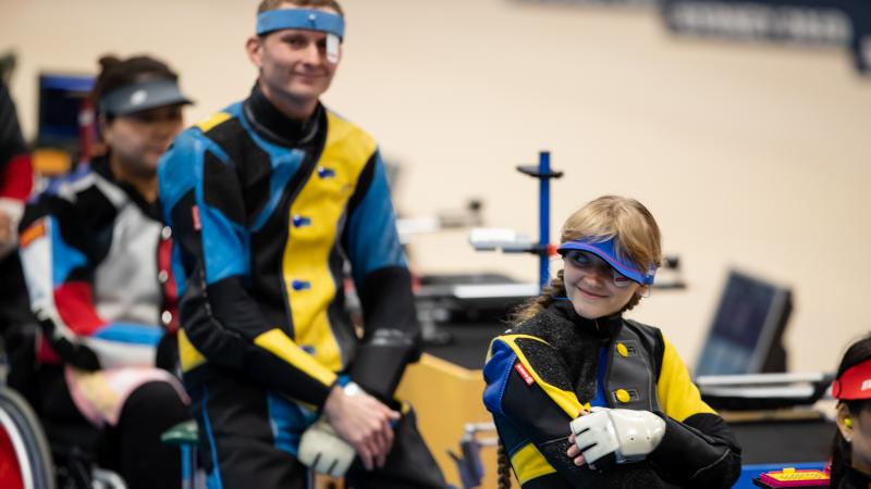 Ukrainian female shooter sits down smiling with male teammate behind also smiling