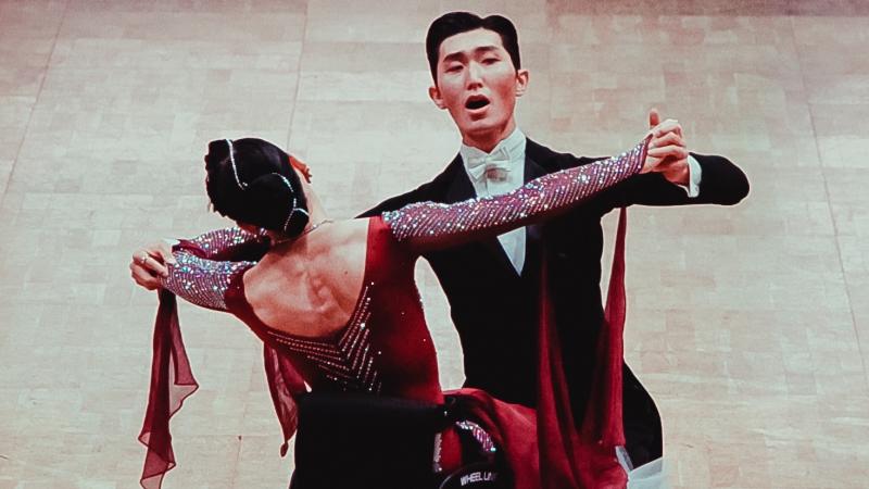 Male standing partner dances with female partner in wheelchair