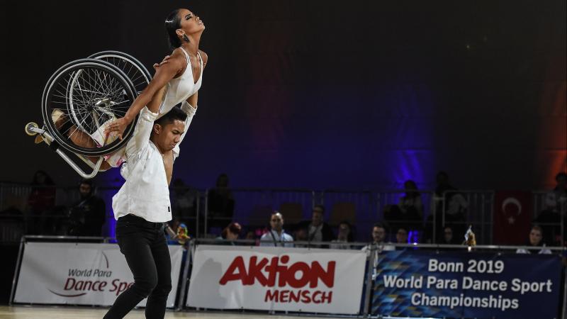 Male standing dancer lifts female wheelchair dancer over his head