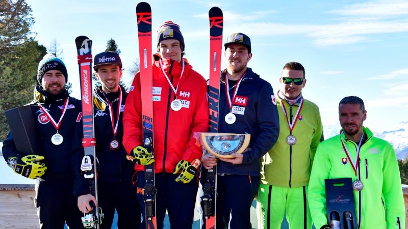 Three vision impaired male skiers with their guides pose on a podium