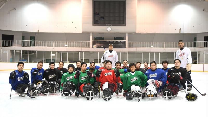 Two men standing on a ice rink behind a group of 20 Para ice hockey players on sledges