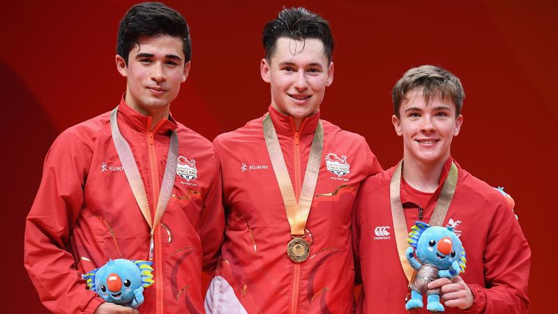 Three British male table tennis players smile on the podium with their medals and mascot prizes