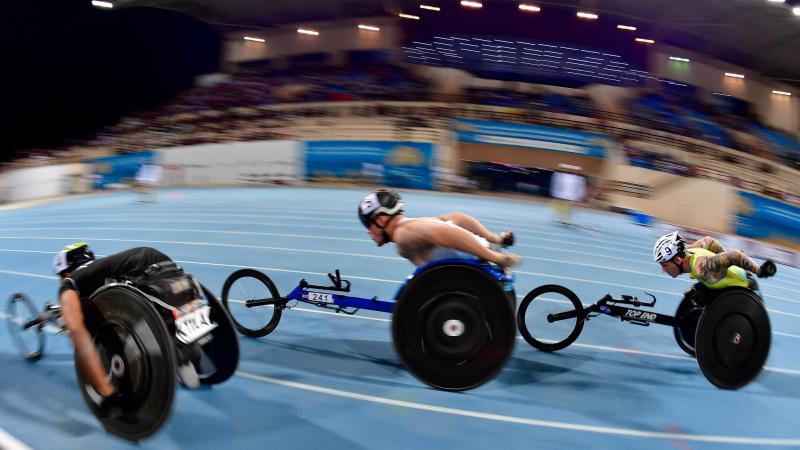 Three men in racing wheelchairs competing in a blue track