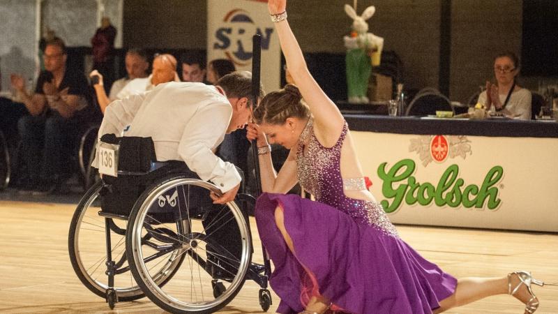 A wheelchair male dancer and a female dancer on a dance floor watched by a group of spectators 