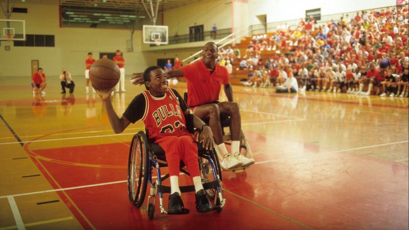 Boy in wheelchair holds basketball while Michael Jordan tries to block