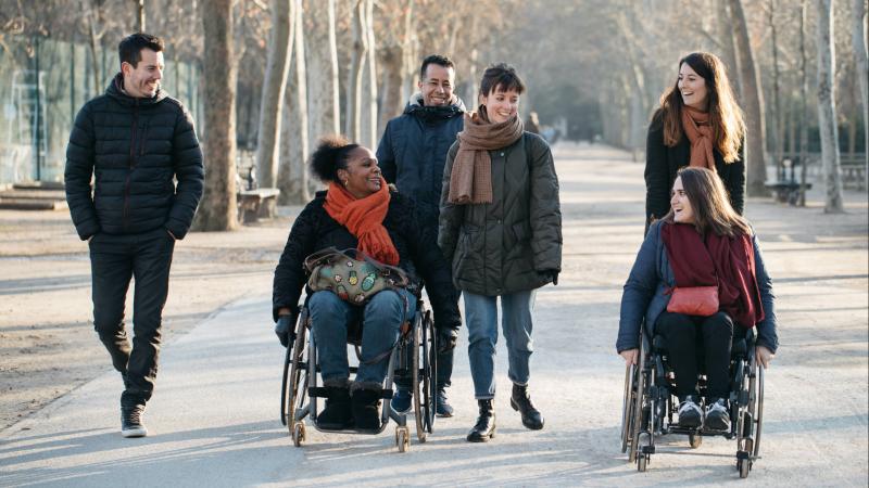 Group of people including two women in a wheelchair walking and smiling in a park