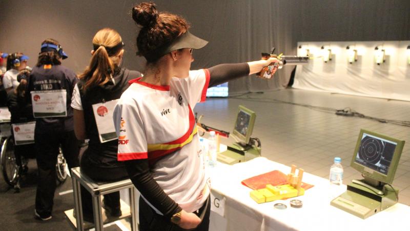 A woman standing and pointing a pistol to a target in an indoor shooting range with four women in the background