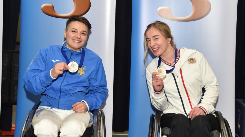 Two female wheelchair fencers pose with their medals on the podium