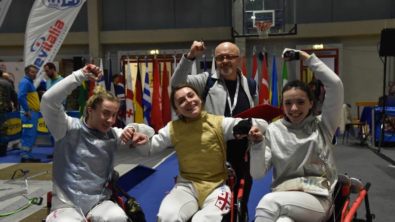 Three female wheelchair fencers with their masks off making a fist in the air