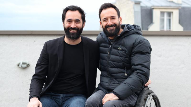 Two French males, one on a wheelchair, take a photo together smiling