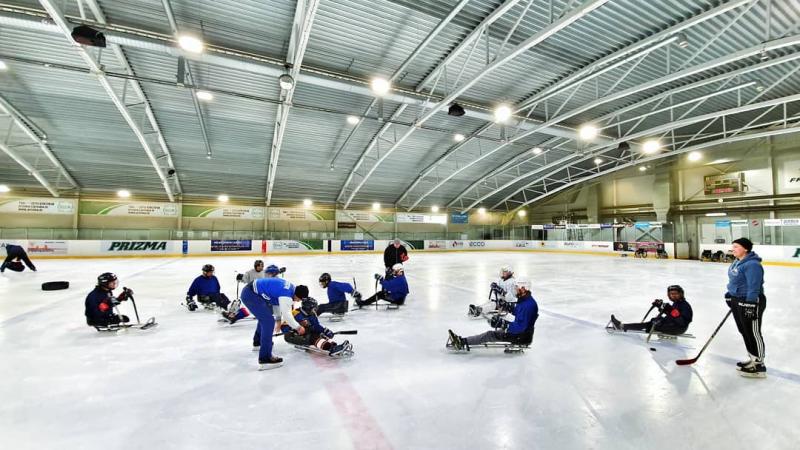 A group of eight persons on sledge hockey receiving instructions from three standing persons on an ice rink
