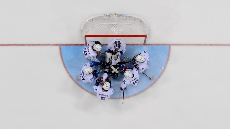 A group of six Para ice hockey players  huddle together in front of the goal