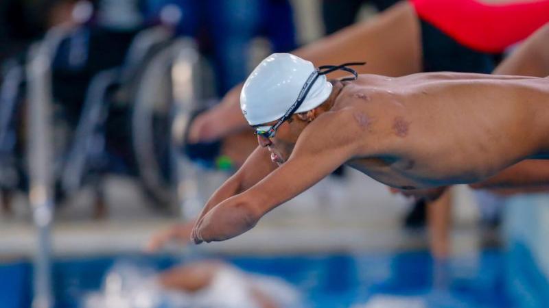 A man without his arms jumping in the swimming pool with another man behind him