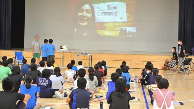 School children in a gym looking at a screen with a woman speaking via video call