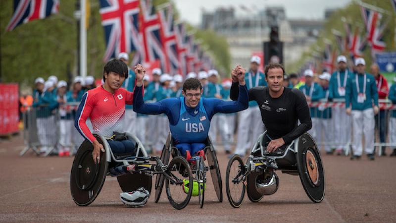 Three men in racing wheelchairs holding hands with the public and flags of the United Kingdom behind them