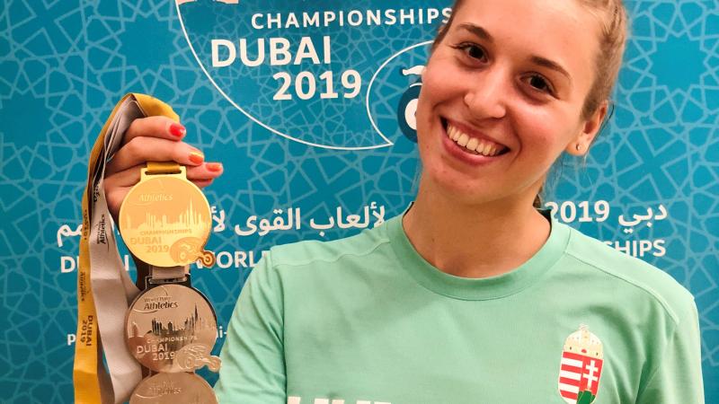 A young woman holding three medals and smiling with a shirt written Hungary