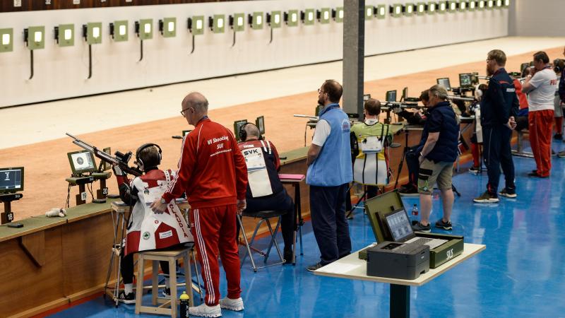 An indoor shooting range with five shooters with one person standing behind each of them