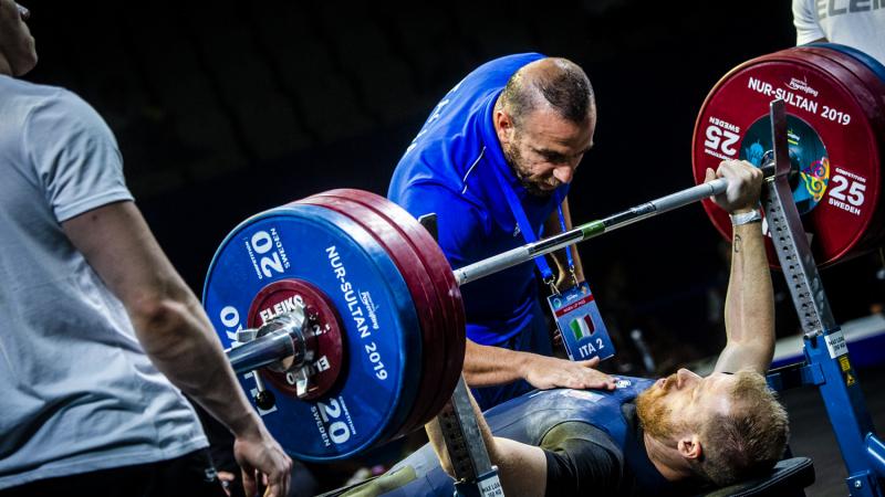 A man on a bench press preparing to lift a bar with three man standing around him