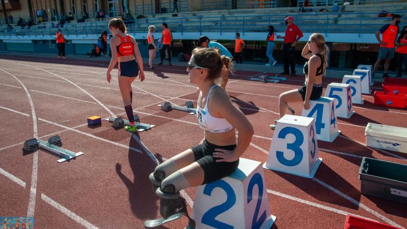 Two women with prosthetic legs sitting on starting blocks on a red athletics track with two other women standing in front of them
