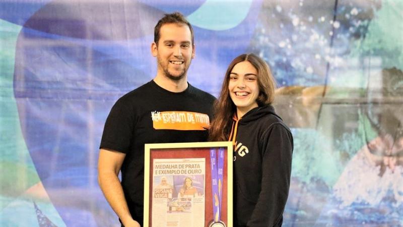 A man and a young woman smiling and holding a frame with a newspaper page and a medal