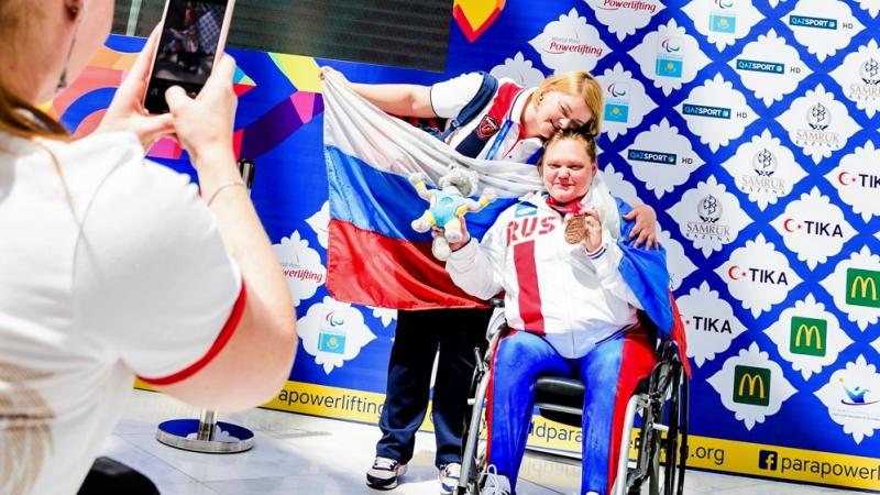 A woman in a wheelchair showing a medal and hugging another woman posing for a photo with the flag of Russia
