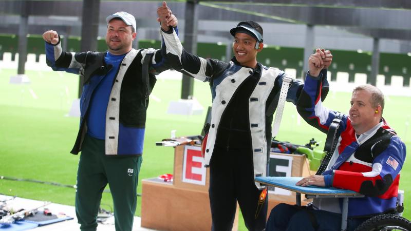 Two men standing and holding hands with a third man in a wheelchair in a shooting range