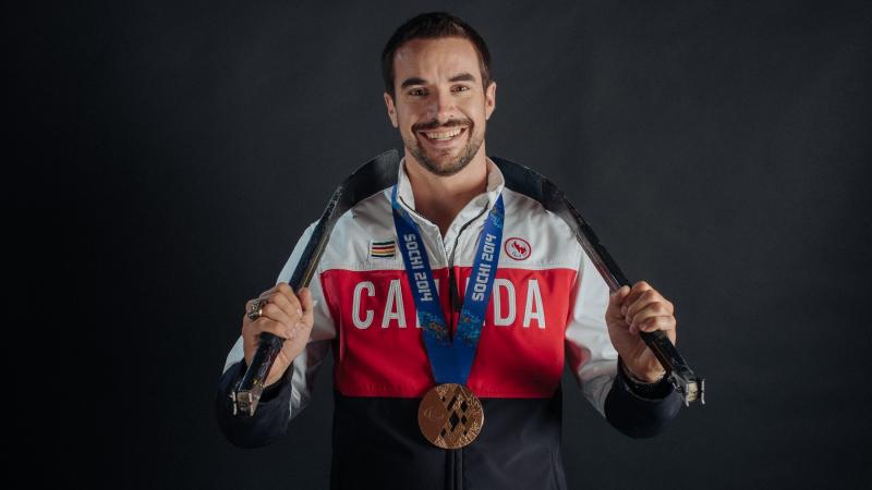 A smiling man with a bronze medal and wearing a Canadian jersey holds hockey sticks behind his neck