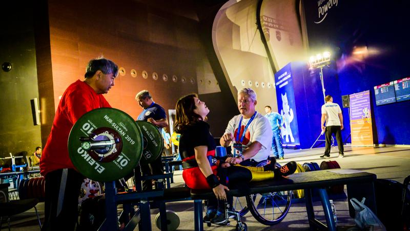 A woman on a bench press talking to a man in a wheelchair right next to her while another man stands near the barbell