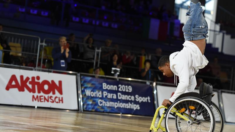 A man without legs upside down holding a wheelchair on a dancefloor 