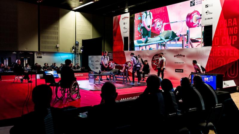 A group of people watching the stage of a Para powerlifting competition with a person competing surrounded by five people