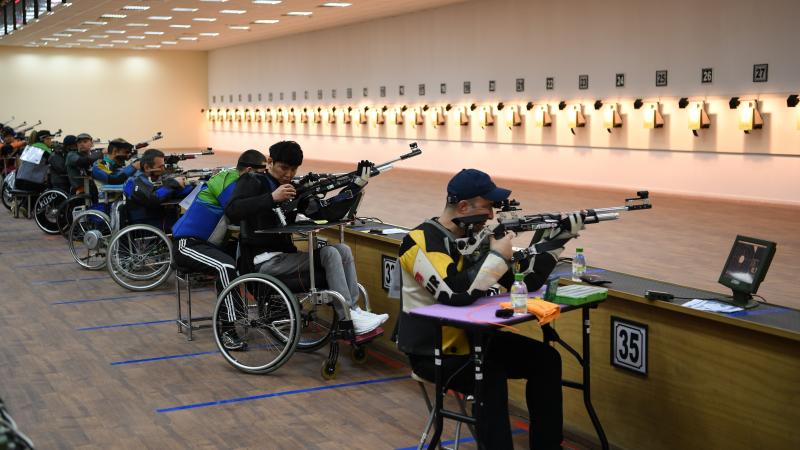 A group of men seated with rifles competing in an indoor shooting range