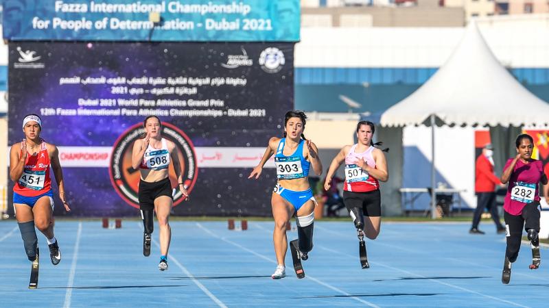 A group of five women with prosthetic legs running on a blue athletics track